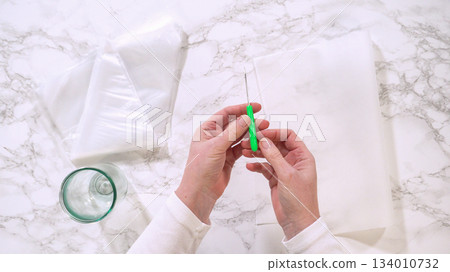 Hands use a green craft knife to cut parchment paper on a marble countertop. A glass bowl and folded parchment are nearby. Hands use a green craft knife to cut parchment paper on a marble countertop. A glass bowl and folded parchment are nearby. 134010732