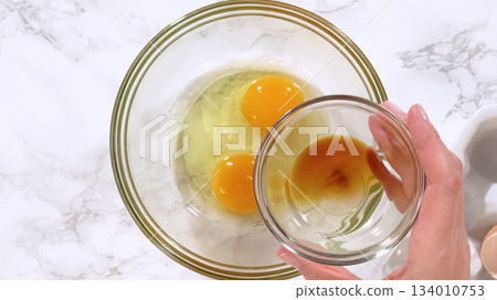 Hands hold a small glass bowl of vanilla extract above a larger mixing bowl containing cracked eggs on a marble countertop. The setup highlights an ingredient preparation step for cookie dough. Hands hold a small glass bowl of vanilla extract above a larger mixing bowl containing cracked eggs on a marble countertop. The setup highlights an ingredient preparation step for cookie dough. 134010753