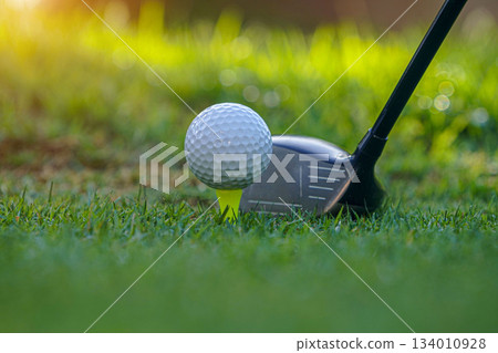 Golf club and golf ball on tee close up in grass field with morning sunshine Golf club and golf ball on tee close up in grass field with morning sunshine 134010928