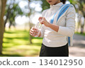 Active Asian adult woman opens water bottle refreshing body after outdoor workout in sunny park promoting hydration and healthy living practices 134012950