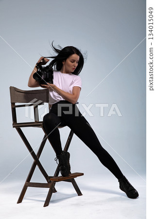 girl sits on a chair with a movie camera in a studio. She appears focused and engaged. The setting suggests a creative atmosphere for filming or practice. 134015049