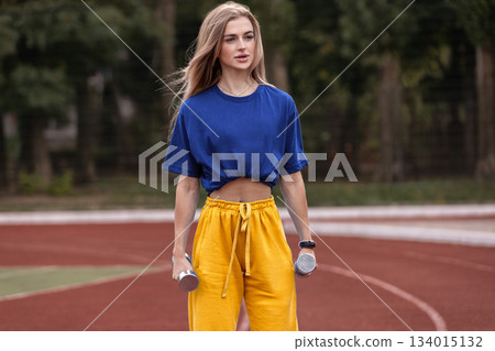 A sporty girl stands on the street holding dumbbells in both hands. She is focused on her workout routine while enjoying her time outdoors in daylight. A sporty girl stands on the street holding dumbbells in both hands. She is focused on her workout routine while enjoying her time outdoors in daylight. 134015132