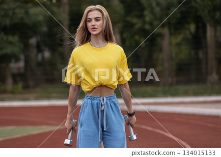 sporty girl stands on a street with dumbbells in her hands. She appears focused and dedicated to her workout. The background shows a sports ground surrounded by trees. sporty girl stands on a street with dumbbells in her hands. She appears focused and dedicated to her workout. The background shows a sports ground surrounded by trees. 134015133
