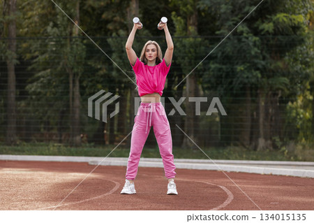 girl stands on a track in the afternoon lifting dumbbells overhead. She wears sporty clothes and focuses on her workout. The trees surround her. girl stands on a track in the afternoon lifting dumbbells overhead. She wears sporty clothes and focuses on her workout. The trees surround her. 134015135
