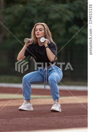 Girl is outdoors on a street holding dumbbells while doing squats. She is engaged in a workout routine focused on strength training. The scene shows outdoor fitness. Girl is outdoors on a street holding dumbbells while doing squats. She is engaged in a workout routine focused on strength training. The scene shows outdoor fitness. 134015138