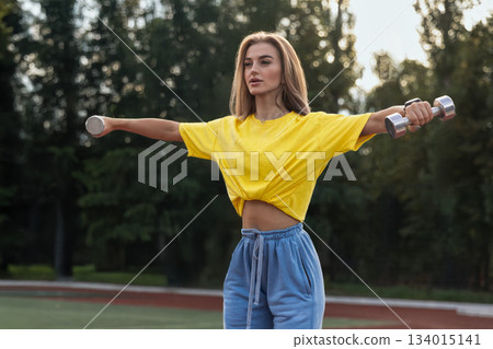 A sporty girl holds dumbbells with arms extended to the side while standing on a street. She is dressed in a yellow top and blue pants. Sunlight shines on her from the late afternoon sky. A sporty girl holds dumbbells with arms extended to the side while standing on a street. She is dressed in a yellow top and blue pants. Sunlight shines on her from the late afternoon sky. 134015141