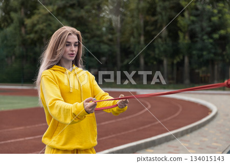 sporty girl practices with elastic bands on a sports ground. She focuses on her workout while surrounded by greenery and track lines. The atmosphere is vibrant and active. sporty girl practices with elastic bands on a sports ground. She focuses on her workout while surrounded by greenery and track lines. The atmosphere is vibrant and active. 134015143