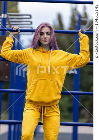 Girl is holding onto a bar in a stadium. She has purple hair and is wearing a yellow hoodie with matching pants. She seems focused and energetic while exercising outdoors. Girl is holding onto a bar in a stadium. She has purple hair and is wearing a yellow hoodie with matching pants. She seems focused and energetic while exercising outdoors. 134015145
