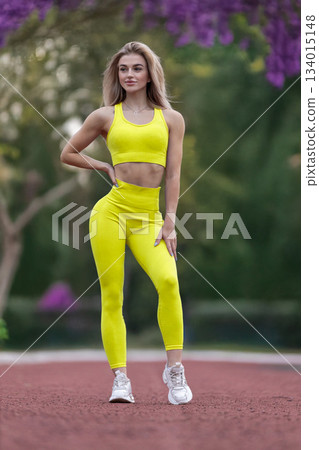 sporty girl stands confidently on the track at the stadium. She wears bright yellow activewear and showcases her athletic build while striking a pose under clear skies. 134015148