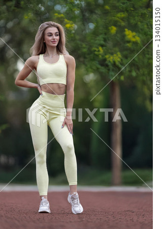 sporty girl stands confidently at the stadium. She wears workout clothes and has long hair. The sun is shining and the atmosphere suggests she is preparing for a training session. 134015150