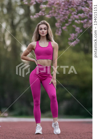 sporty girl stands on the track at a stadium showing her strength and focus. She wears bright workout clothes and has a confident stance. It is a sunny afternoon. 134015151