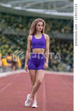 sporty girl walks confidently along the track at the stadium. She is dressed in a purple outfit and is getting ready for her training session. sporty girl walks confidently along the track at the stadium. She is dressed in a purple outfit and is getting ready for her training session. 134015152