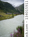 Vertical shot of alpine village scenery with glacial river flowing through and massive glacier above 134015911