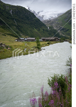 Vertical shot of alpine village scenery with glacial river flowing through and massive glacier above Vertical shot of alpine village scenery with glacial river flowing through and massive glacier above 134015911