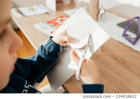 Hands holding scissors while cutting a shape from white paper on a wooden table. Natural light highlights a calm, creative moment, suitable for themes of crafting, learning, hobbies and home education Hands holding scissors while cutting a shape from white paper on a wooden table. Natural light highlights a calm, creative moment, suitable for themes of crafting, learning, hobbies and home education 134016652