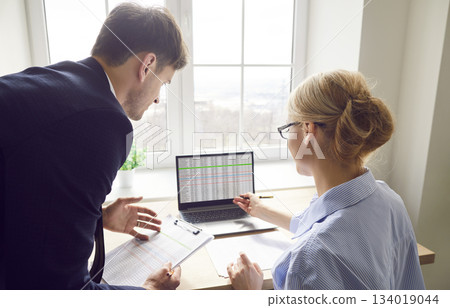 Two company employees sitting with laptop computer in office looking at monitor screen. 134019044