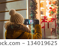 Woman tourist in knitted hat takes photographs of New Year's festive city decor on her smartphone. Christmas market, night lights. View from the back. 134019625