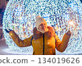Young woman in knitted hat holds sparkler in her hands. Night festive street, shiny city decorations for Christmas. 134019626