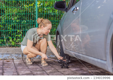 Woman lifting a car with a jack on the roadside, trying to change a tire while handling tools on her own. Automotive repair, independence and problem-solving concept, representing confidence Woman lifting a car with a jack on the roadside, trying to change a tire while handling tools on her own. Automotive repair, independence and problem-solving concept, representing confidence 134019996