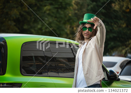 Caucasian adult man 30s wearing green hat and novelty sunglasses standing near car outdoors. St patricks day celebration, lifestyle portrait with festive mood. Caucasian adult man 30s wearing green hat and novelty sunglasses standing near car outdoors. St patricks day celebration, lifestyle portrait with festive mood. 134021372