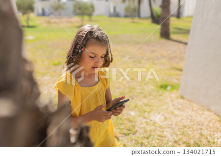 Girl 7 years old in yellow dress talking on video call on phone on lawn with palm trees in summer day 134021715