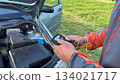 A Caucasian male mechanic in an orange and gray jacket adjusting a glow plug tester with yellow wires near the engine bay of a silver car on a grassy roadside 134021717