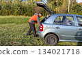 A Caucasian male mechanic in an orange and gray jacket retrieving tools from the open trunk of a silver car parked on a grassy roadside in daylight 134021718