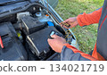 A Caucasian male mechanic in an orange and gray jacket adjusting a glow plug tester while holding glow plugs near the open engine bay of a silver car 134021719