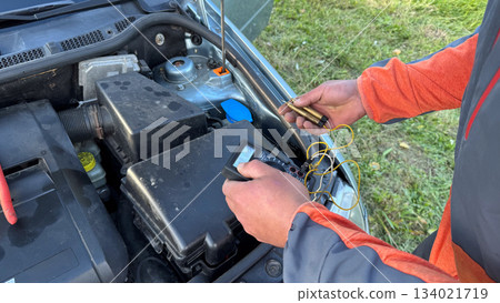 A Caucasian male mechanic in an orange and gray jacket adjusting a glow plug tester while holding glow plugs near the open engine bay of a silver car A Caucasian male mechanic in an orange and gray jacket adjusting a glow plug tester while holding glow plugs near the open engine bay of a silver car 134021719