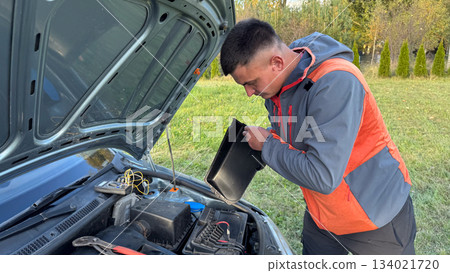 A Caucasian male mechanic in an orange and gray jacket opening the battery compartment of a silver car with the hood raised on a grassy roadside 134021720