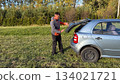 A Caucasian male mechanic in an orange and gray jacket opening the trunk of a silver car from the side view while standing on green grass under afternoon sunlight 134021721