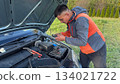 A Caucasian male mechanic in an orange and gray jacket connecting a glow plug tester to the car battery under the open hood of a silver vehicle on a grassy roadside 134021722