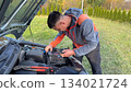 A Caucasian male mechanic in an orange and gray jacket using a glow plug tester connected to the battery of a silver car with the hood open on a grassy roadside 134021724