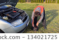 A Caucasian male mechanic in an orange and gray jacket bending down to take tools from a black bag next to a silver car with the hood open on a grassy roadside 134021725