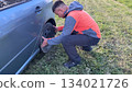 A Caucasian male mechanic in an orange and gray jacket inspecting the left rear wheel of a silver car while crouching on a grassy roadside 134021726