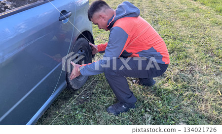 A Caucasian male mechanic in an orange and gray jacket inspecting the left rear wheel of a silver car while crouching on a grassy roadside A Caucasian male mechanic in an orange and gray jacket inspecting the left rear wheel of a silver car while crouching on a grassy roadside 134021726
