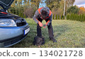 A Caucasian male mechanic in an orange and gray jacket checking the glow plug tester device while standing next to an open tool bag near a silver car with the hood open 134021728