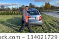 A Caucasian male mechanic in a gray and orange jacket opening the trunk of a silver car parked on a grassy roadside under a clear blue sky 134021729
