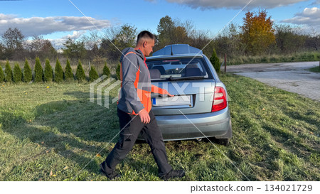 A Caucasian male mechanic in a gray and orange jacket opening the trunk of a silver car parked on a grassy roadside under a clear blue sky A Caucasian male mechanic in a gray and orange jacket opening the trunk of a silver car parked on a grassy roadside under a clear blue sky 134021729
