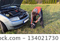 A Caucasian male mechanic in an orange and gray jacket retrieving a glow plug tester from a black tool bag next to a silver car with the hood open on a grassy roadside 134021730
