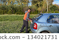 A Caucasian male mechanic in an orange and gray jacket closing the trunk of a silver car while standing on a grassy roadside in sunny weather 134021731