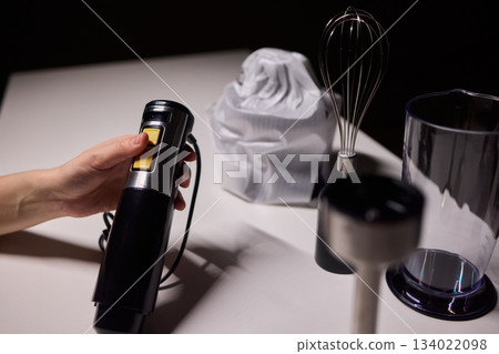Black handheld immersion blender in professional kitchen setting, Kitchen expert demonstrating use of handheld immersion blender beside measuring tools and ingredients 134022098