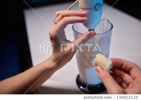 Hands prepare creamy banana smoothie, Electric blender mixes banana and milk for smoothie, Closeup of hands combining banana and milk to create refreshing breakfast drink 134022110