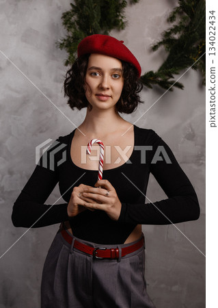 Caucasian woman holding candy cane in festive room with grey wall and green garland Caucasian woman holding candy cane in festive room with grey wall and green garland 134022434