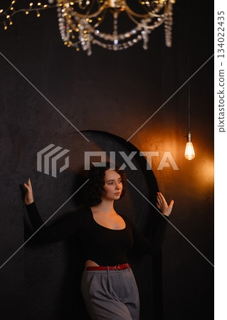 Caucasian woman posing by dark textured wall with warm lightbulb in moody room Caucasian woman posing by dark textured wall with warm lightbulb in moody room 134022435