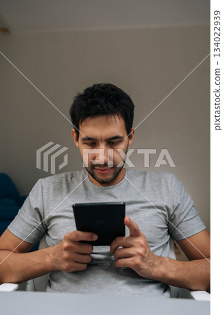 Vertical portrait of young adult man holding black e-reader, comfortably reading and studying digital content at home, focused on screen in relaxed modern indoor setting. Vertical portrait of young adult man holding black e-reader, comfortably reading and studying digital content at home, focused on screen in relaxed modern indoor setting. 134022939