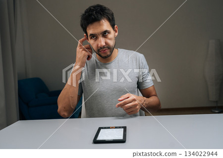 Pensive young man using electronic reader at home, engaging with digital content for study or leisure, illustrating modern portable reading and learning technology. 134022944