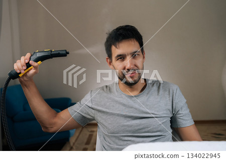 Portrait of cheerful man having fun with steam cleaner, showing humorous attitude during household chores performance, enjoying cleaning process with playful approach. Portrait of cheerful man having fun with steam cleaner, showing humorous attitude during household chores performance, enjoying cleaning process with playful approach. 134022945