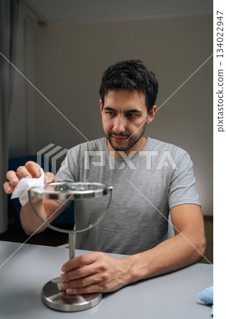 Vertical portrait of man using blue microfiber cloth to carefully clean round tabletop mirror, performing household chore with focus on personal hygiene and maintaining clean environment. Vertical portrait of man using blue microfiber cloth to carefully clean round tabletop mirror, performing household chore with focus on personal hygiene and maintaining clean environment. 134022947