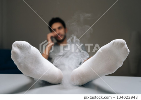Selective focus of man relaxing talking on phone with feet up on table, white socks emitting white smoke representing bad smell or stench, signifying strong foot odor and unpleasant hygiene issues. 134022948